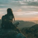 Solo Traveler Sunset - woman sitting on rock carrying black backpack